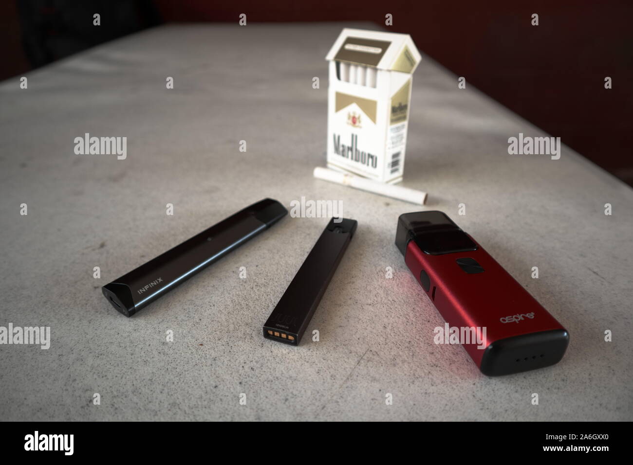 3 vapes juul, aspirent à la brise, smok infinix avec un paquet de cigarettes de Marlboro et une cigarette à l'extérieur sur une table isolée, texturé Blanc Banque D'Images