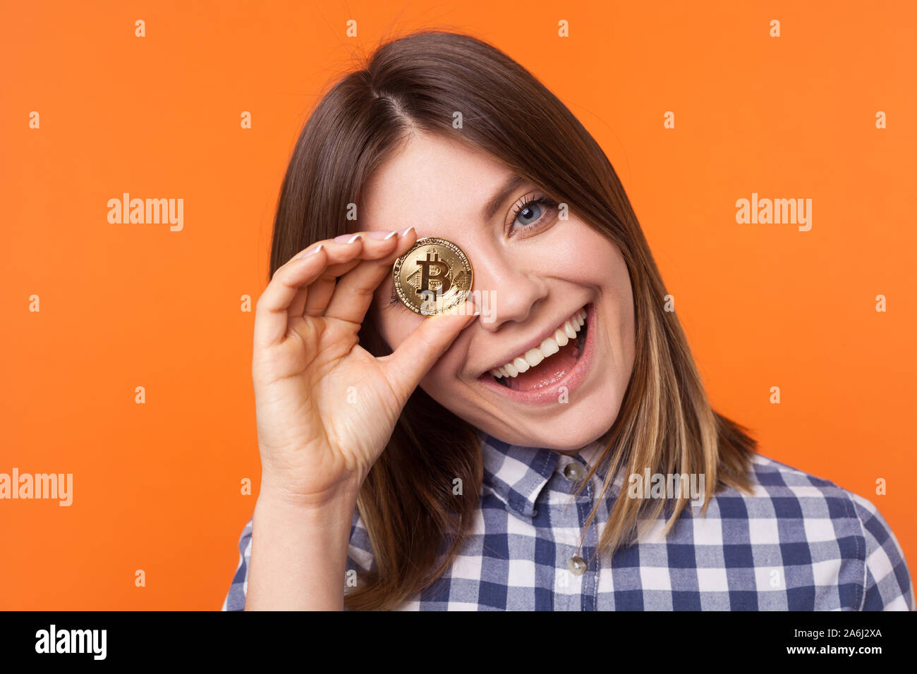 Closeup portrait of playful brunette woman avec charmant sourire portant chemise à carreaux couvrant un oeil d'or, cryptocurrency avec bitcoin. Banque D'Images
