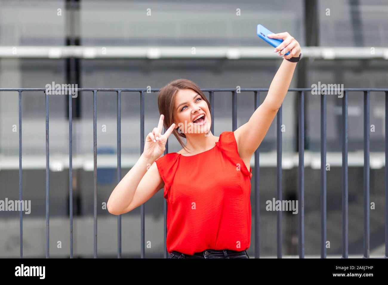Portrait de jeune femme brune fantaisie dans l'élégant quartier de chemise rouge en tenant sur smartphone selfies et montrant la victoire ou geste de paix, de célébrer la réussite. mak Banque D'Images