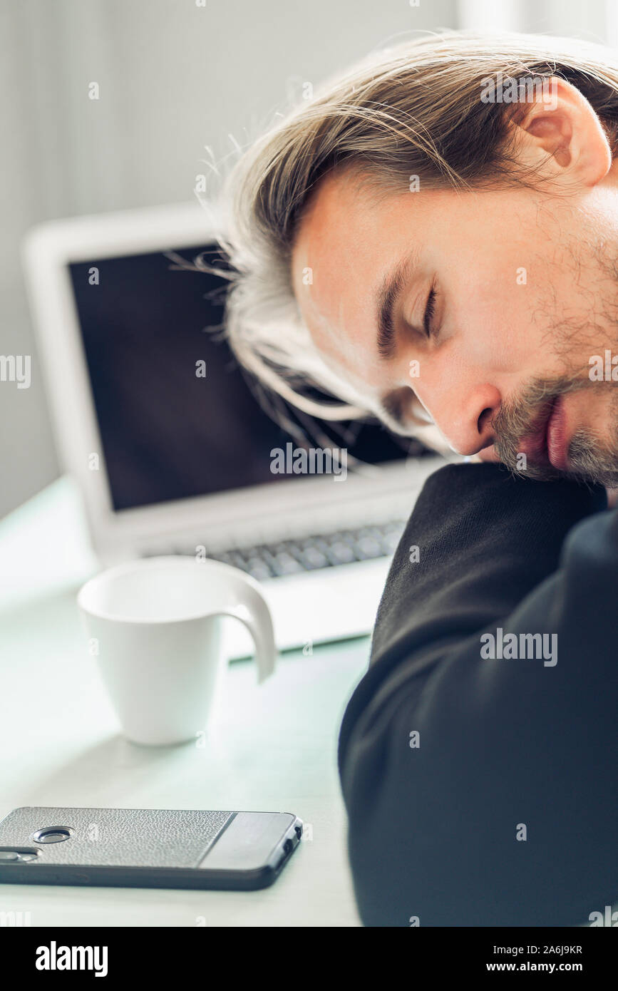 Beau jeune homme dormir sur pile de livres au bureau. Le café, le téléphone et un ordinateur portable sur un bureau à côté de lui. Banque D'Images