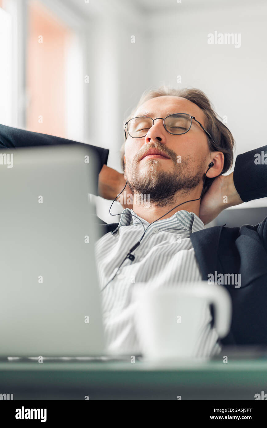Jeune homme essaie de se détendre et de dormir tout en écoutant de la musique au travail. Tasse à café et un ordinateur visible au premier plan. Banque D'Images