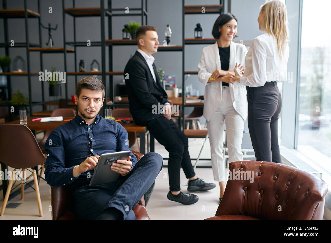 Beau mec assis sur le fauteuil avec tablette, regardant la caméra pendant que ses trois collègues ayant l'amusement dans l'arrière-plan de la photo. Banque D'Images
