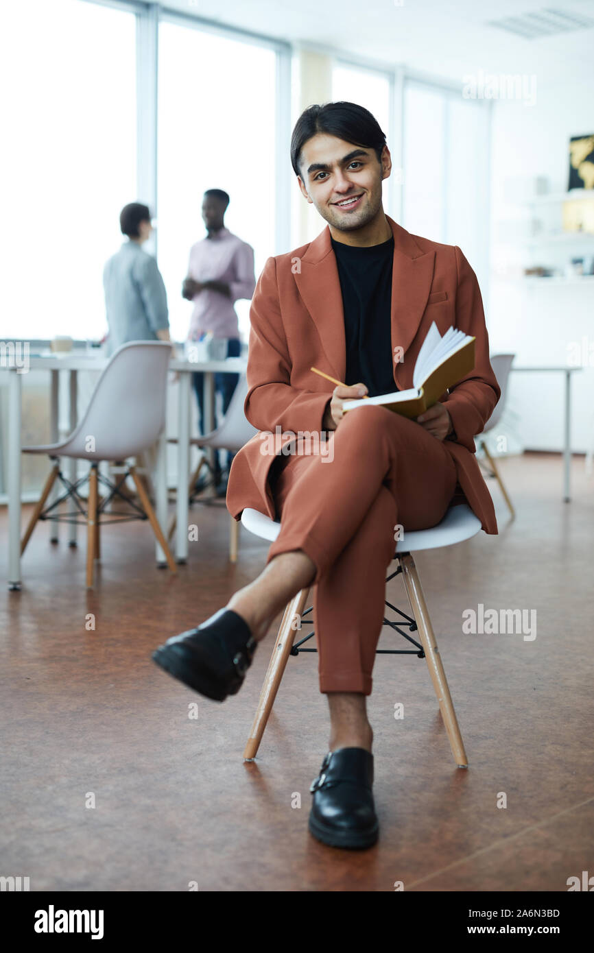 Portrait de jeune homme du Moyen-Orient smiling at camera while sitting on chair in modern office Banque D'Images