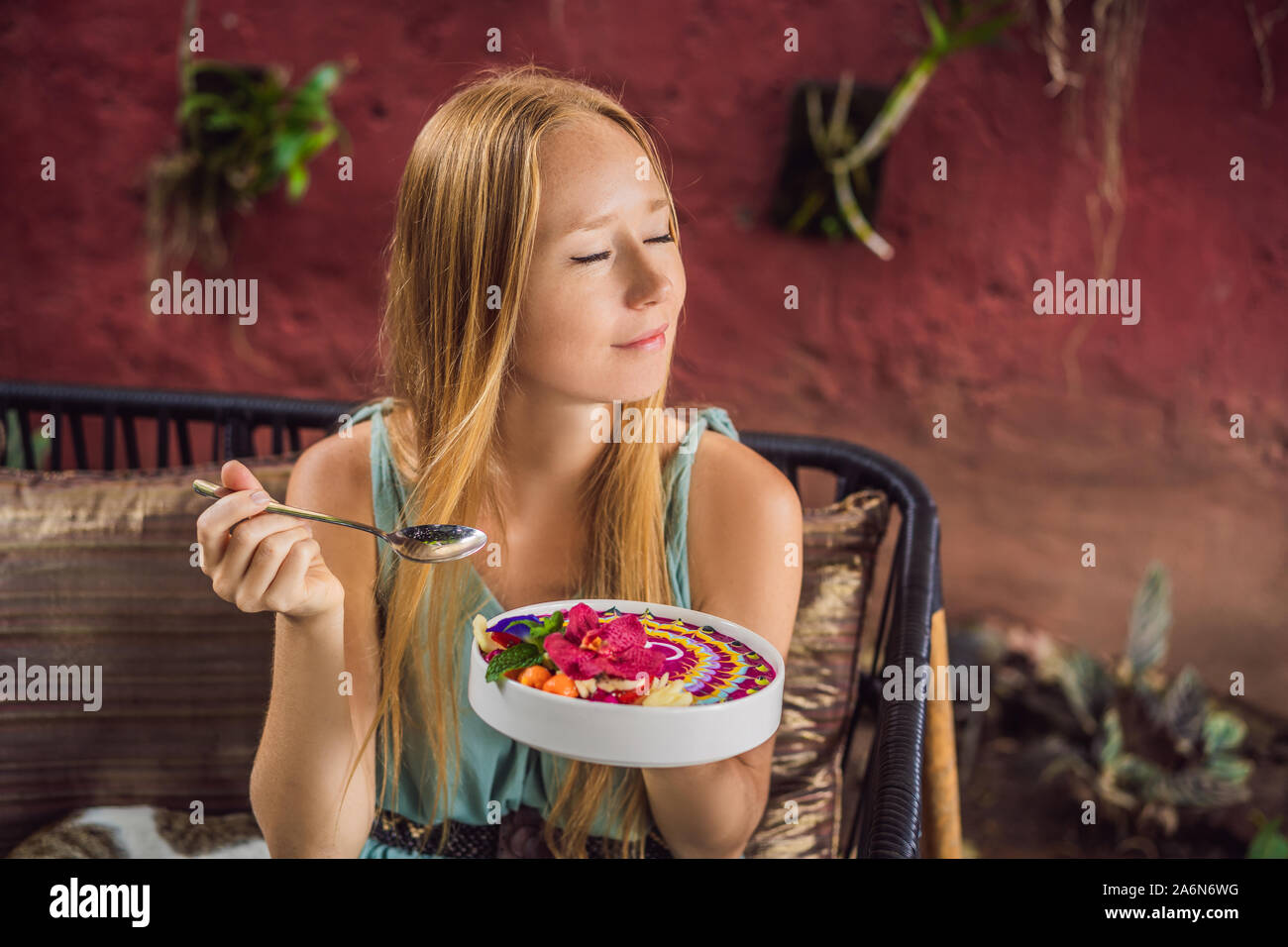 Jeune femme ayant un petit déjeuner méditerranéen assis à son canapé et mange le petit déjeuner tropical smoothie sain, bol de fruits tropicaux, décoré Banque D'Images