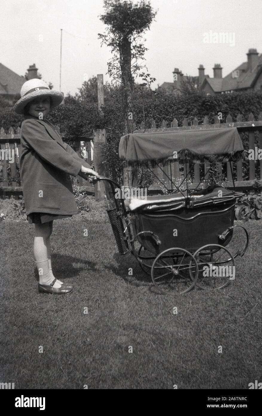 Circa 1930s, historique, à l'extérieur dans le jardin arrière, une jeune fille portant une veste d'école et chapeau de paille debout avec ses poupées poussette, Angleterre Royaume-Uni. Banque D'Images