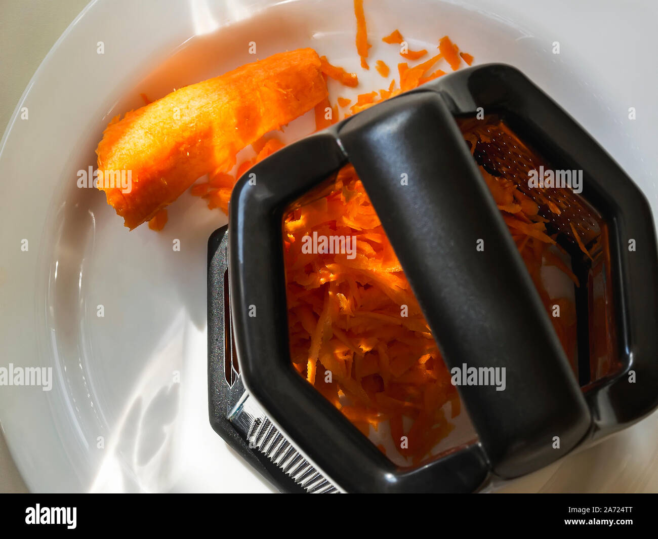 Râpe à légumes en métal avec une poignée en plastique noir et une carotte râpée crue dans une assiette blanche. Des légumes frais pour une alimentation saine, la nourriture végétarienne. Banque D'Images