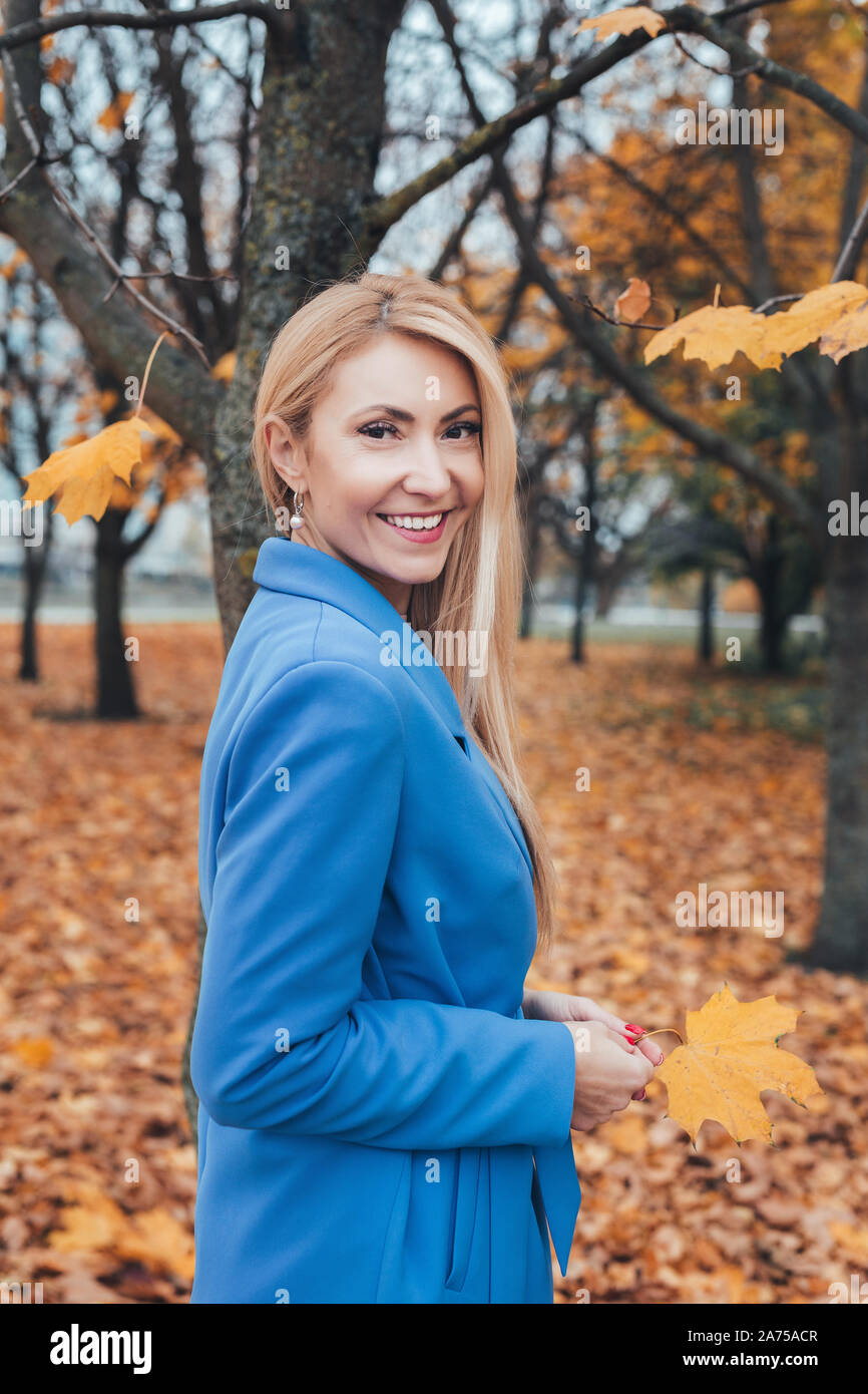 Smiling blonde mature woman in blue coat looking at camera in autumn park Banque D'Images