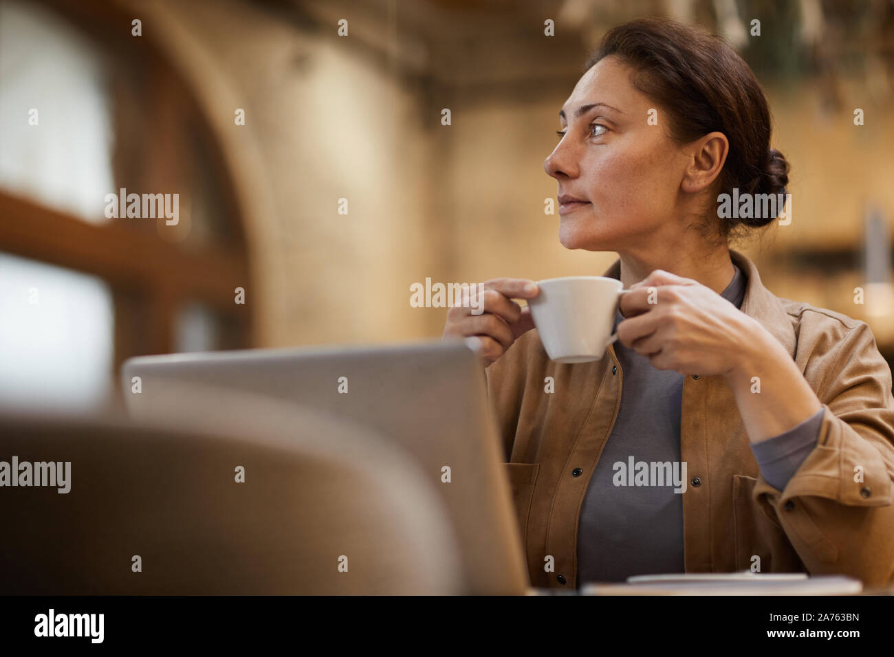 Mature Woman holding tasse de café et à l'écart avec la vue tout en restant assis et pensif working on laptop Banque D'Images