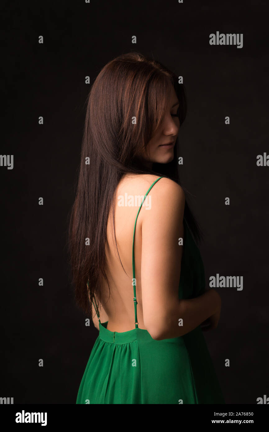 Une belle brune aux cheveux longs dans une robe verte avec un dos nu se dresse sur un fond sombre. Pensive, romantique et naturel sexy. Concept pour les cheveux c Banque D'Images