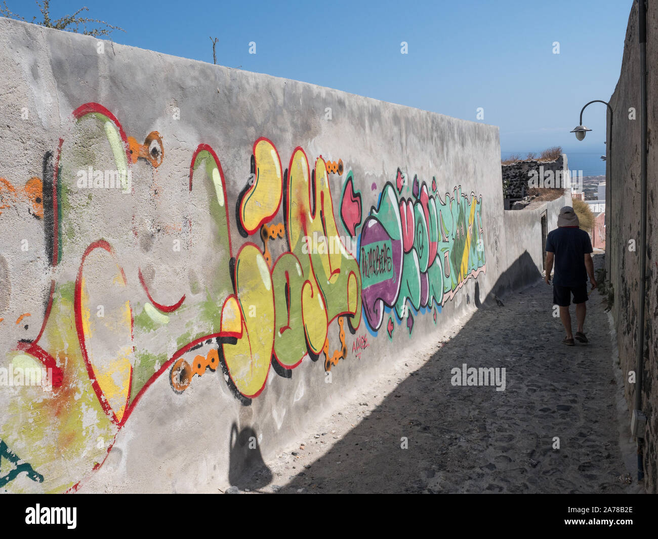 Une vue en perspective d'un mur de béton avec des graffitis lumineux de l'ombre et horizon de mer avec arrière-plan touristique mâle en passant par Fira Santorini Banque D'Images