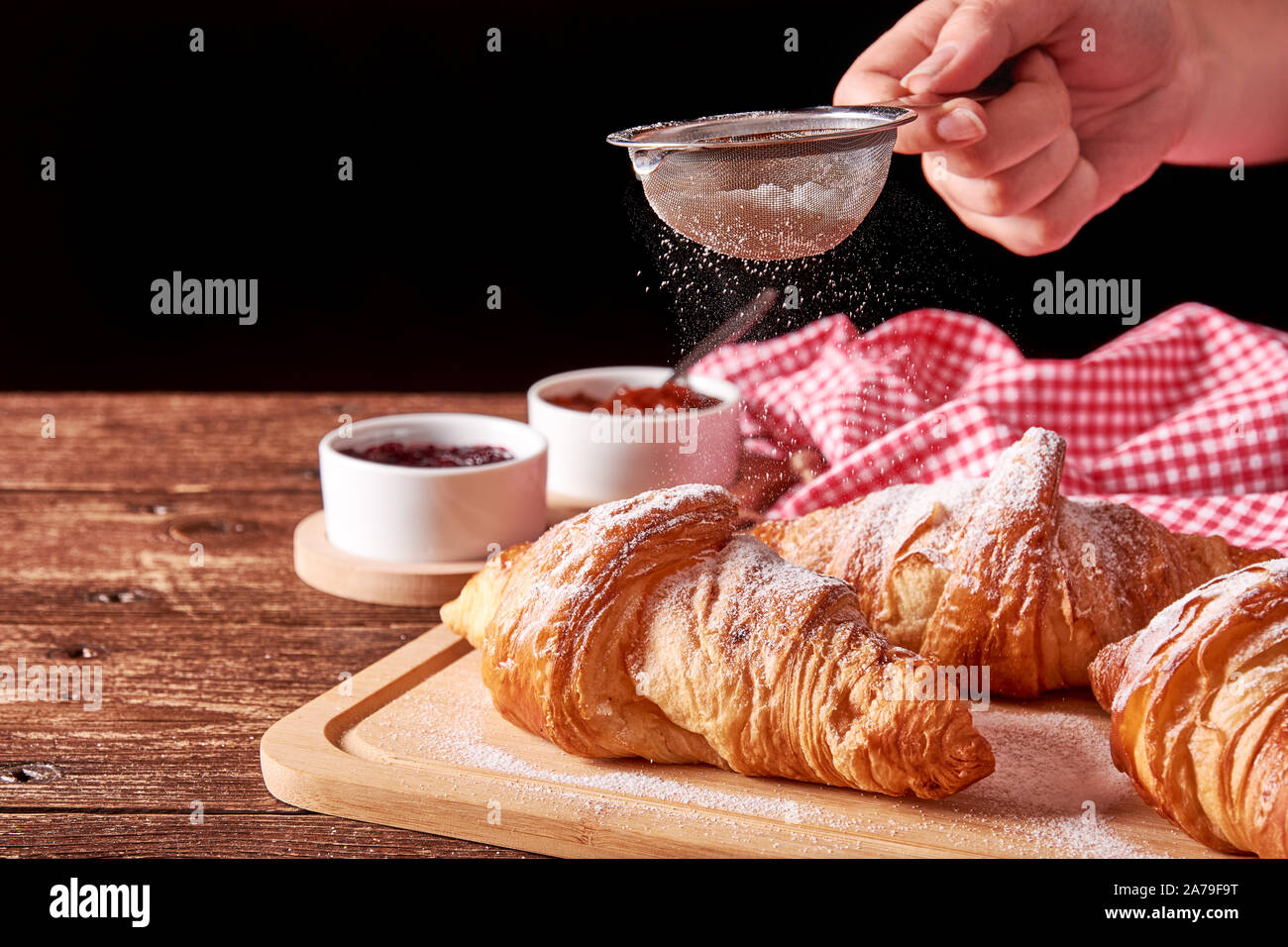 La vie encore sur de vieux tapis en bois avec trois croissants et de la confiture de fruits confits à la pétanque. Main tenant un tamis de sucre sur un fond noir. Banque D'Images