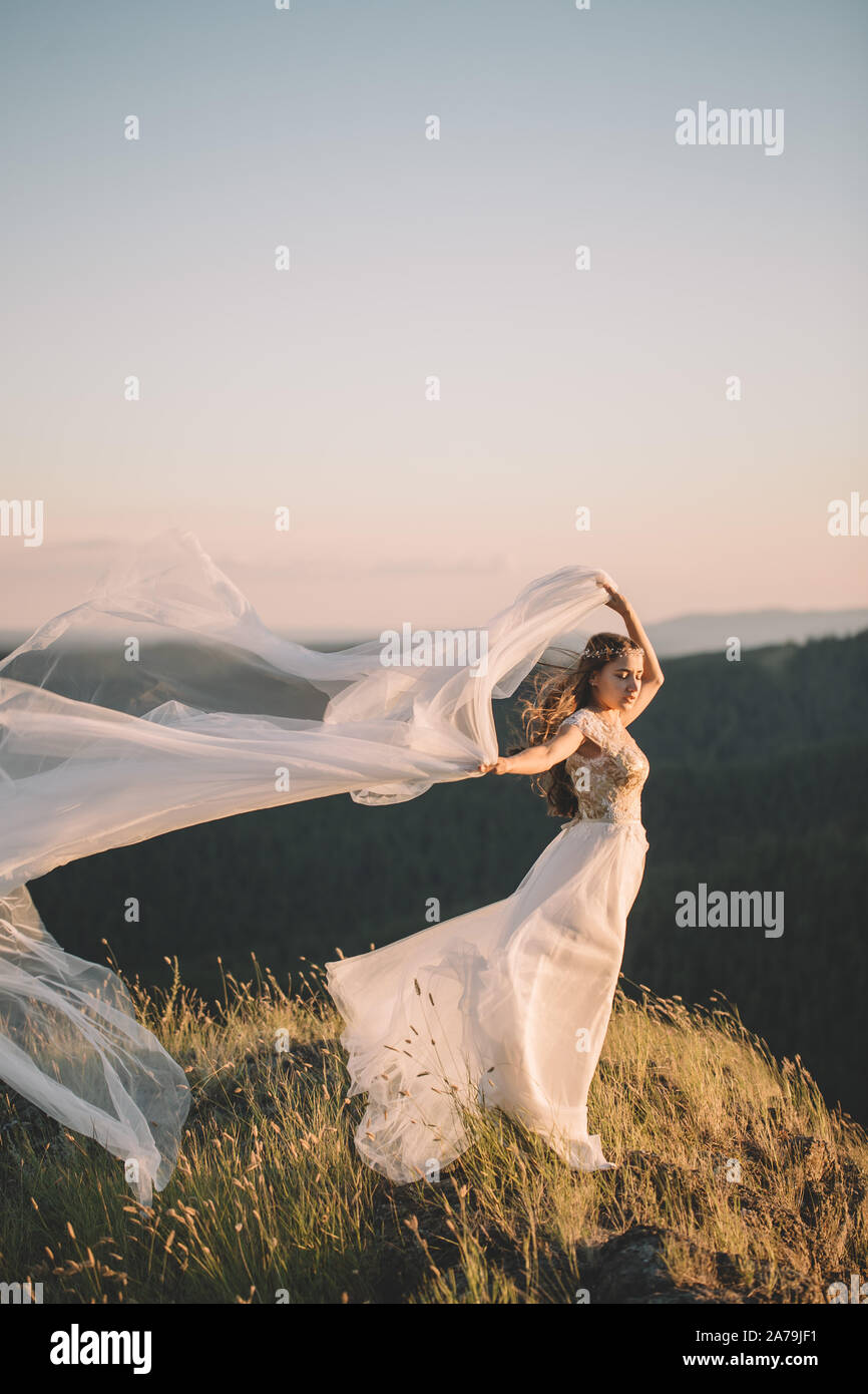 Belle jeune femme brune dans un voile blanc le curling dans le vent et dans  une robe blanche en haut d'une haute montagne en été à l'extérieur. Fine  art, weddin Photo Stock -