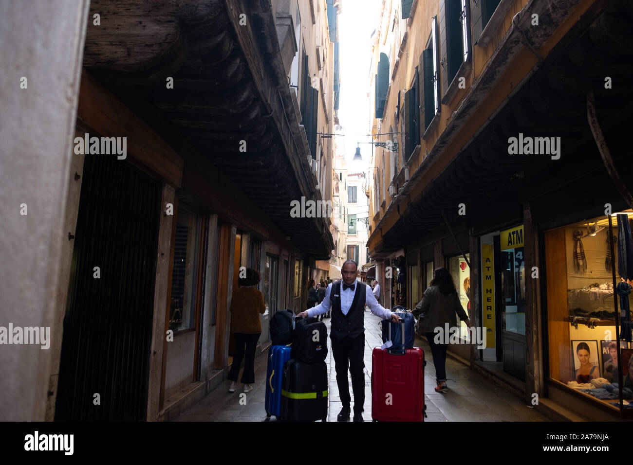 Un hôtel à porter l'uniforme noir et blanc tirant deux chargé assurance trollies vers l'observateur dans une étroite, surplombée, rue commerçante de Venise. Banque D'Images