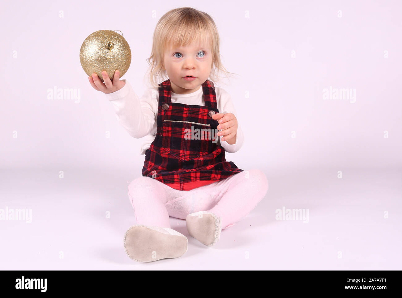 Petite blonde bébé fille avec de grands yeux bleus en robe rouge jouer avec Boule de Noël,assis sur le plancher, fond blanc.Portrait isolé Banque D'Images