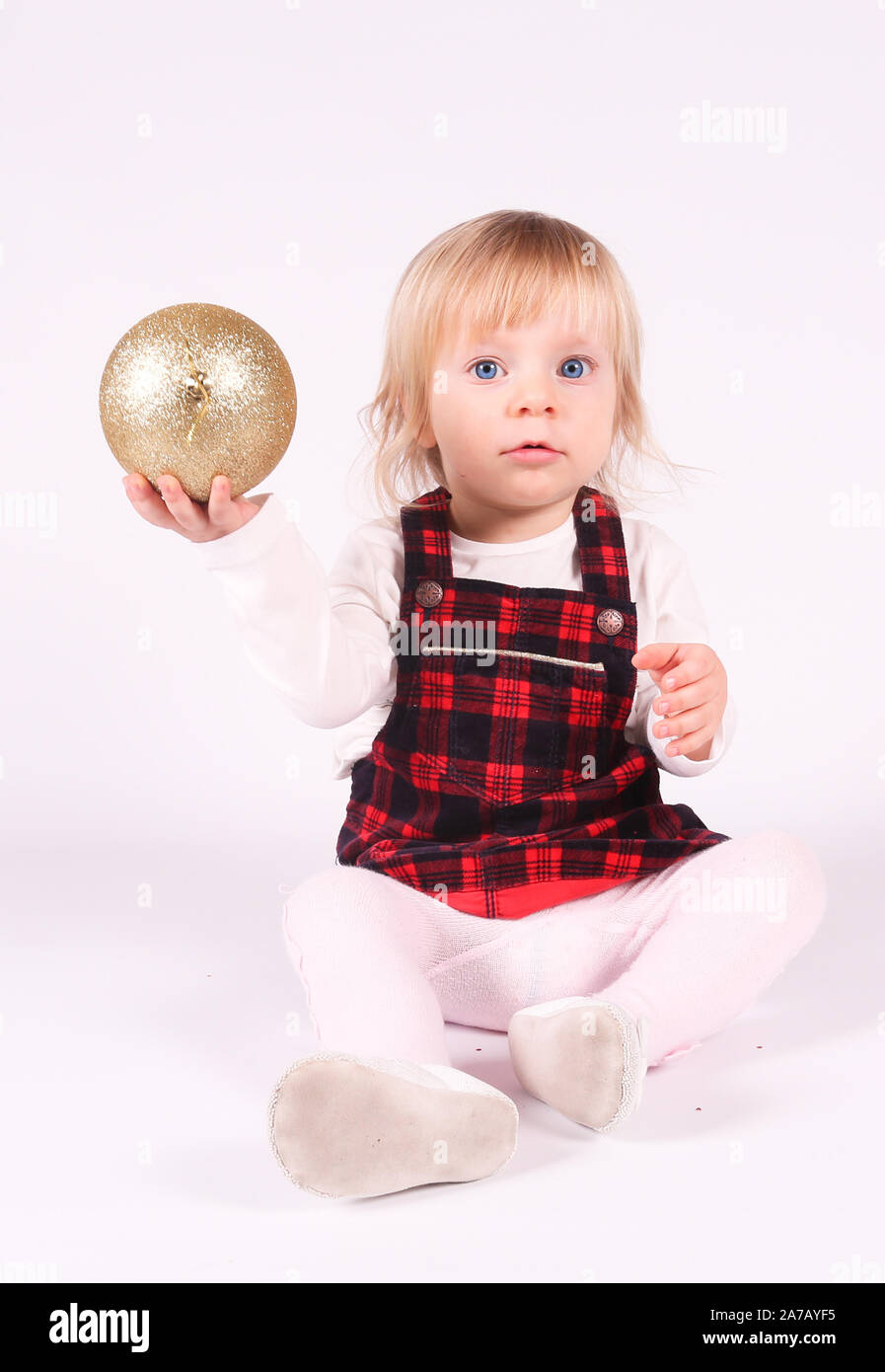 Petite blonde bébé fille avec de grands yeux bleus en robe rouge jouer avec Boule de Noël,assis sur le plancher, fond blanc.Portrait isolé Banque D'Images
