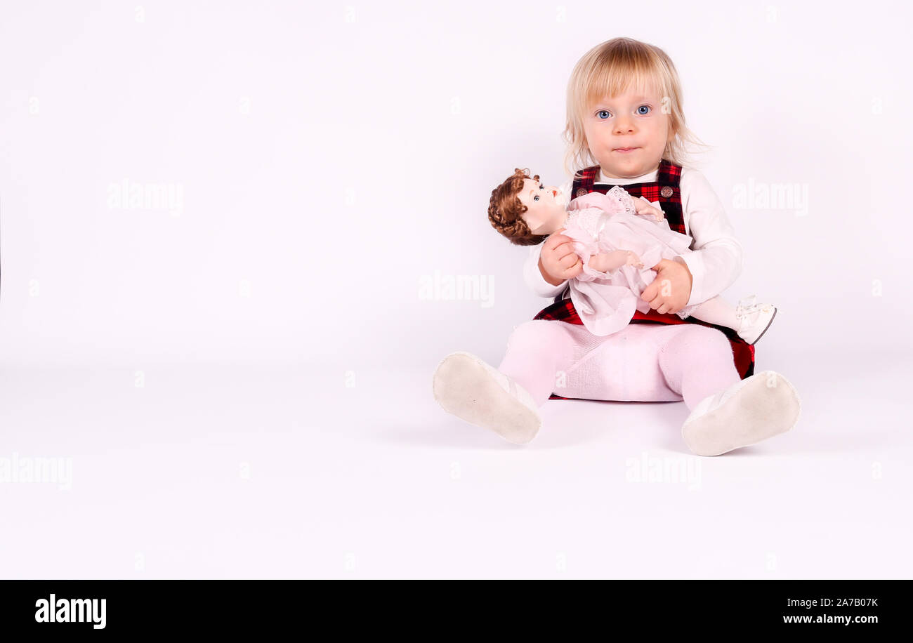 Petite blonde bébé fille avec de grands yeux bleus en robe rouge jouer avec l'ancienne poupée. Assis sur le plancher, fond blanc. Portrait isolé Banque D'Images