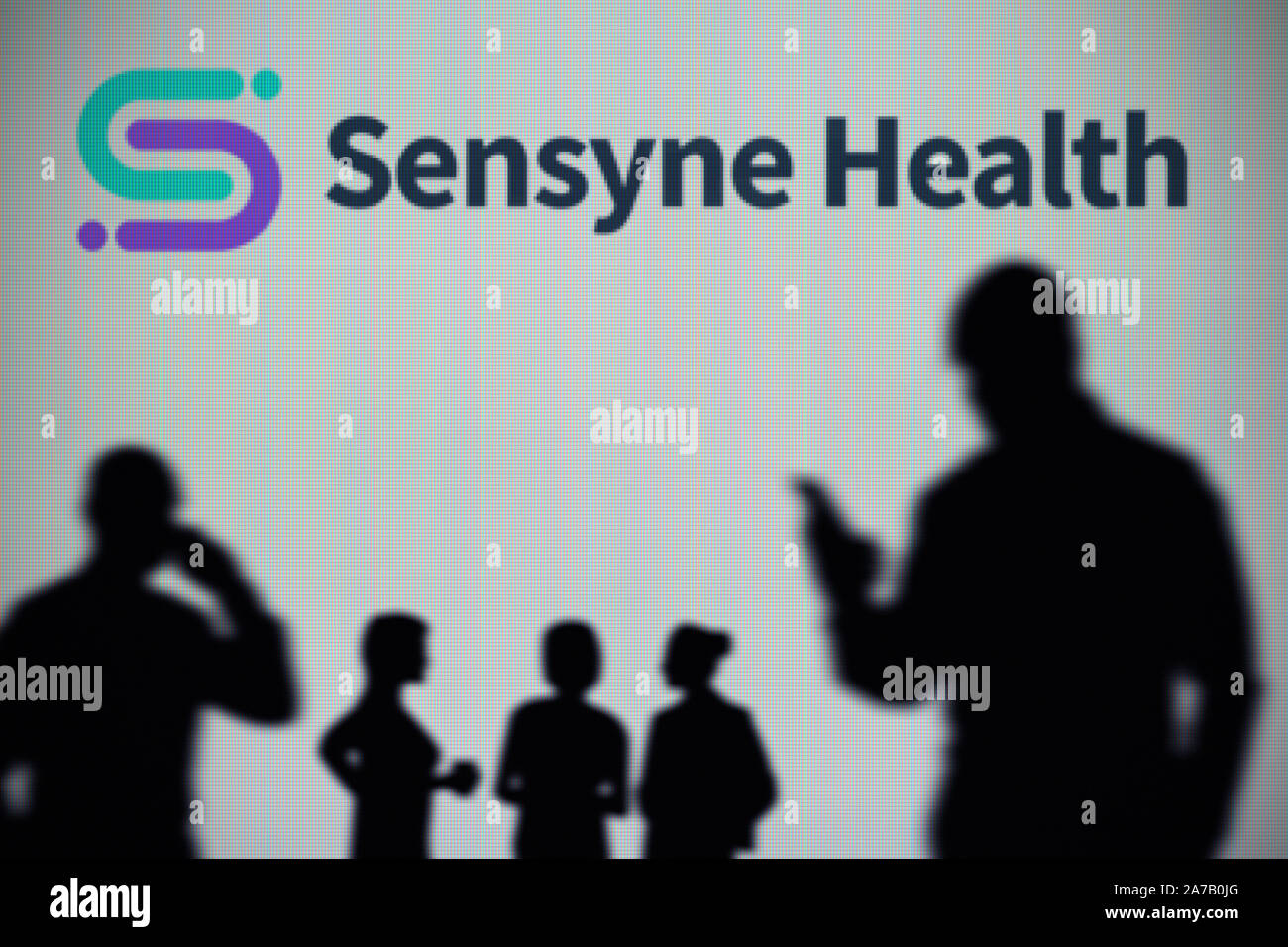 Le Sensyne logo Santé est visible sur un écran LED à l'arrière-plan tandis qu'une silhouette personne utilise un smartphone (usage éditorial uniquement) Banque D'Images