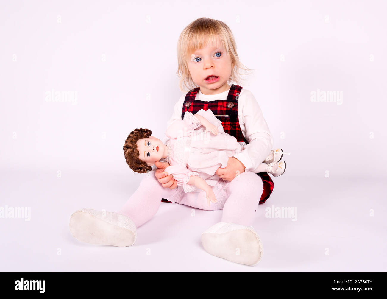 Petite blonde bébé fille avec de grands yeux bleus en robe rouge jouer avec l'ancienne poupée. Assis sur le plancher, fond blanc. Portrait isolé Banque D'Images