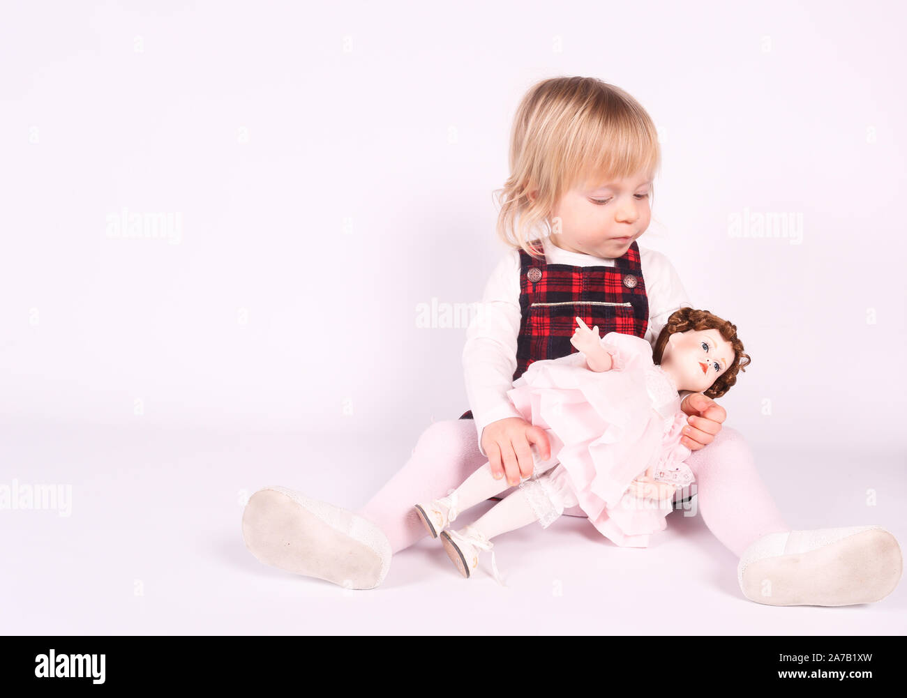 Petite blonde bébé fille avec de grands yeux bleus en robe rouge jouer avec l'ancienne poupée. Assis sur le plancher, fond blanc. Portrait isolé Banque D'Images