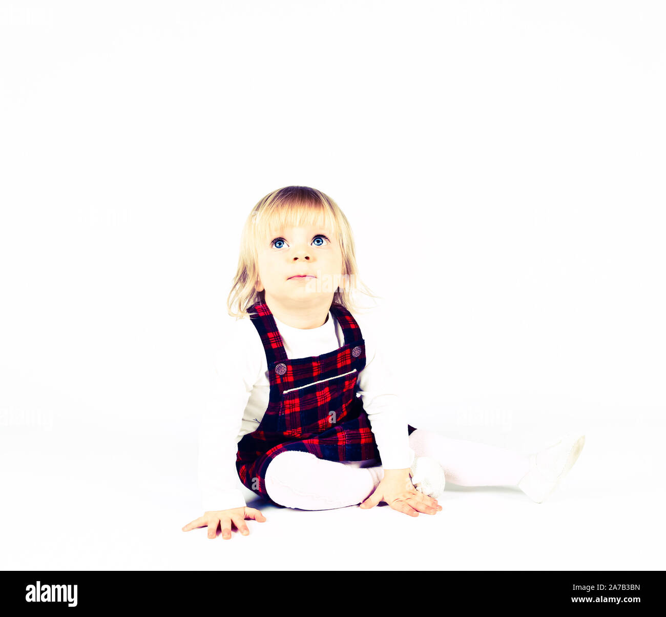 Petite blonde bébé fille avec de grands yeux bleus en robe rouge assise sur le sol, fond blanc. Portrait isolé Banque D'Images