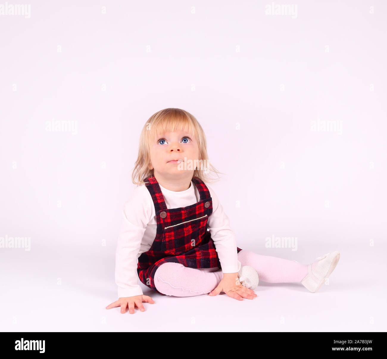 Petite blonde bébé fille avec de grands yeux bleus en robe rouge assise sur le sol, fond blanc. Portrait isolé Banque D'Images