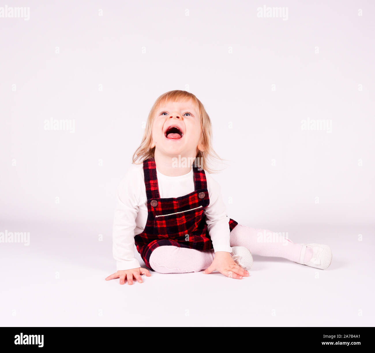 Petite blonde bébé fille avec de grands yeux bleus en robe rouge assise sur le sol, fond blanc. Portrait isolé Banque D'Images