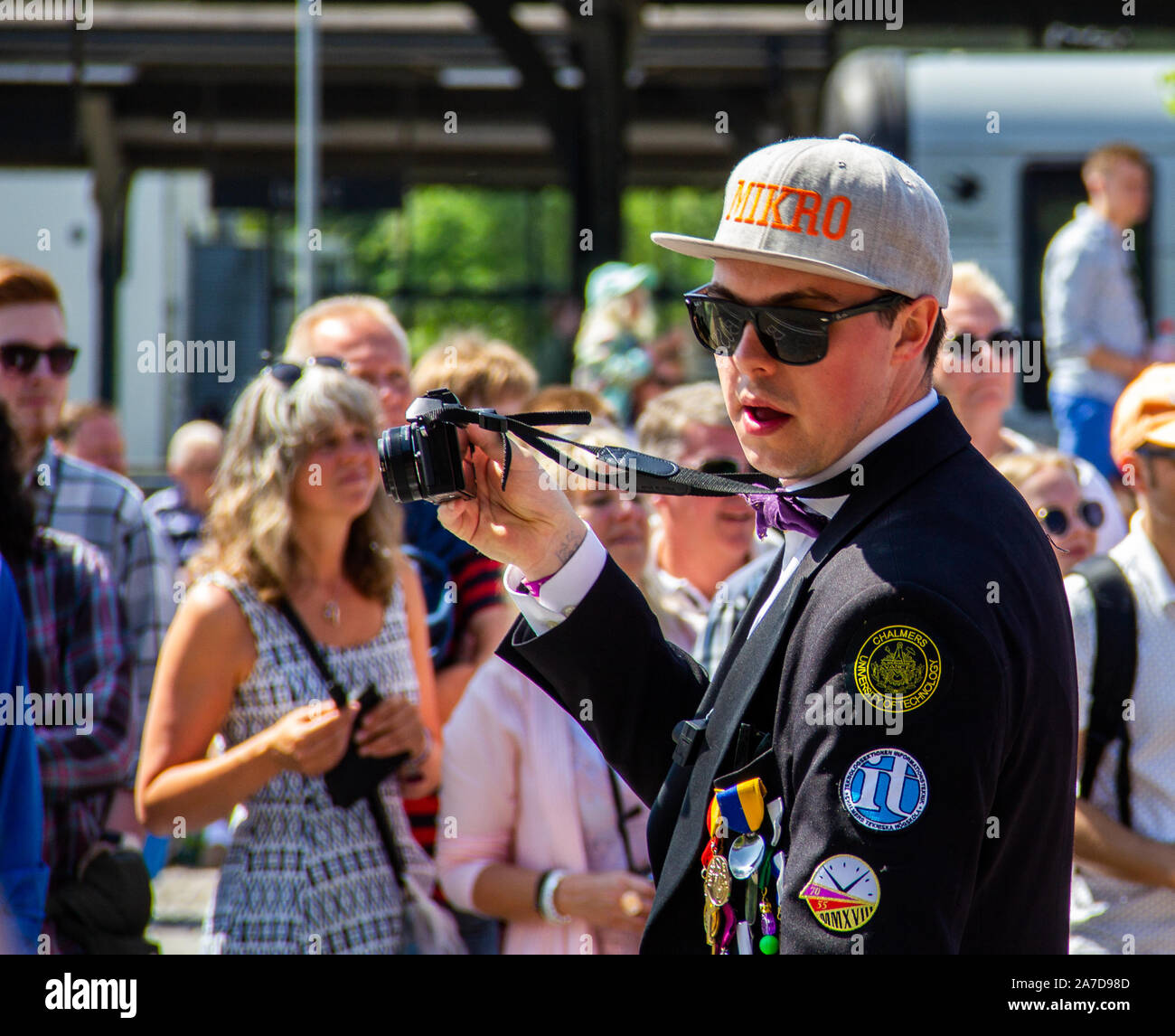 Un jeune homme participant à la parade Lundakarnevalen tout en prenant des photos en tant que photographe. Il porte un chapeau, des lunettes et un costume avec marquage Banque D'Images