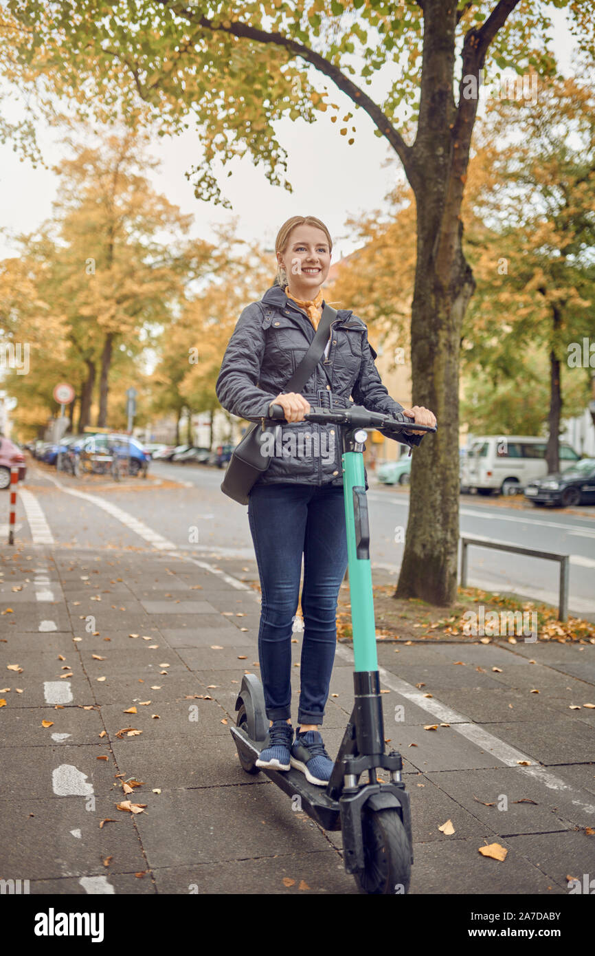Young happy blonde woman riding scooter électrique dans la ville, souriant à la caméra, à l'automne Banque D'Images