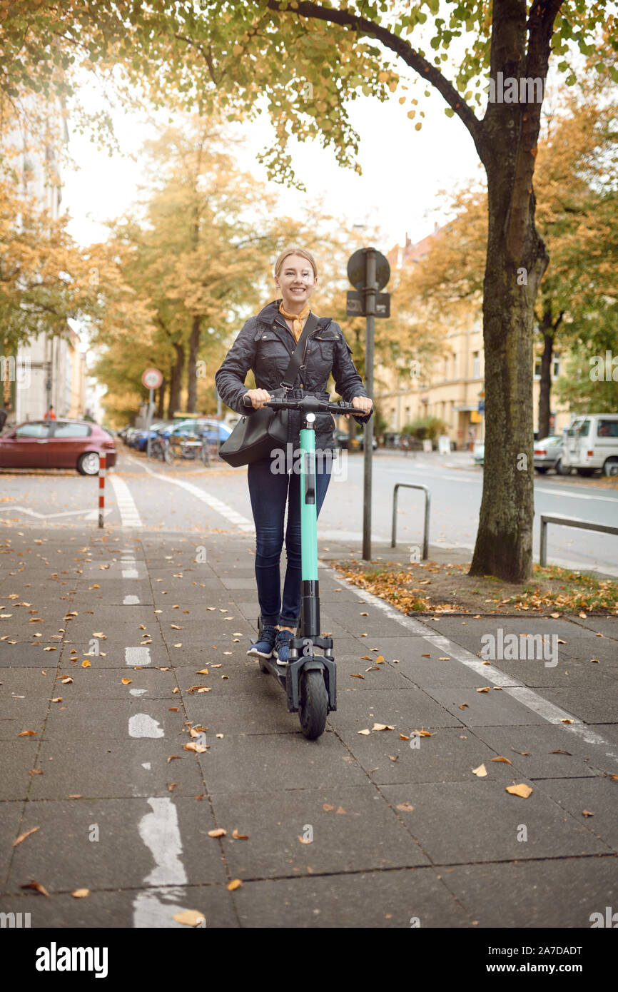 Young happy blonde woman riding scooter électrique dans la ville, souriant à la caméra, à l'automne Banque D'Images