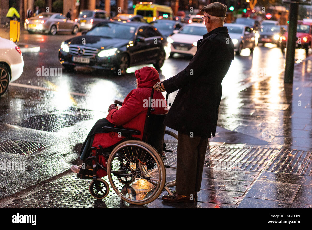 Un homme âgé attend à un passage pour piétons avec son fauteuil roulant avec sur un soir de pluie dans le quartier londonien de Kings Cross Banque D'Images
