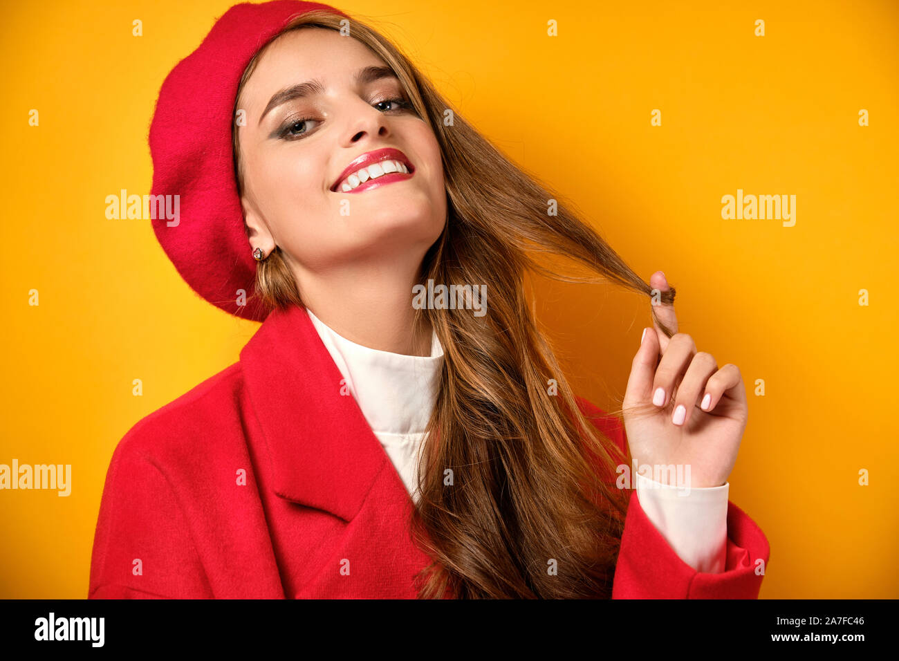 Une fille avec des boucles, le rouge à lèvres rouge dans un manteau rouge et un béret est debout sur un fond jaune, virevoltant une boucle avec son doigt. Banque D'Images