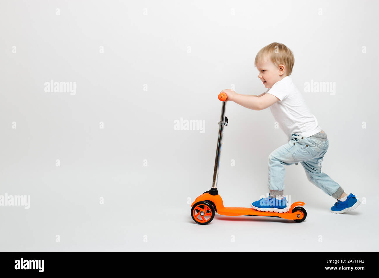 Heureux tout-petit enfant dans une orange en jeans scooter sur un fond blanc dans le studio. La publicité pour les jouets pour enfants avec l'espace pour le texte. Banque D'Images