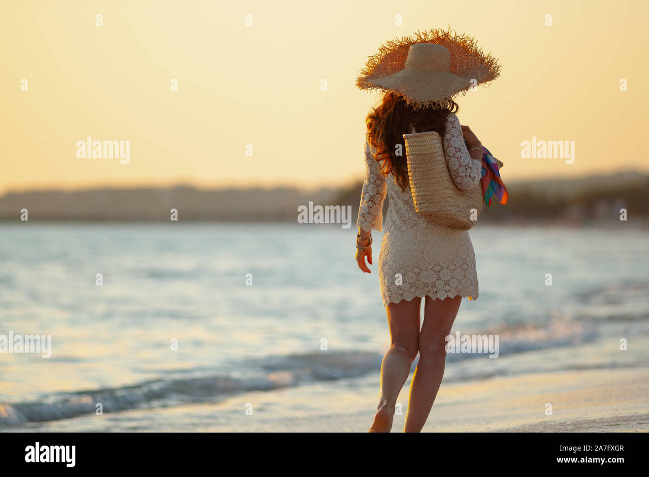 Vus de derrière, l'âge moyen élégant femme en robe blanche et chapeau de paille sur la plage au coucher du soleil la marche. Banque D'Images