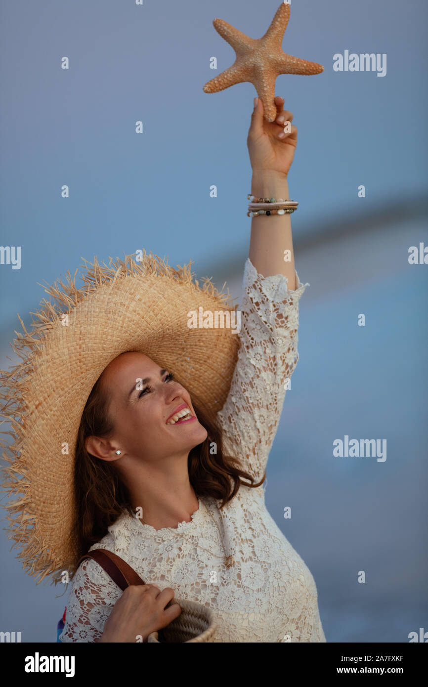 Moderne souriant de 40 ans, femme en robe blanche et chapeau de paille sur la plage au coucher du soleil holding starfish. Banque D'Images
