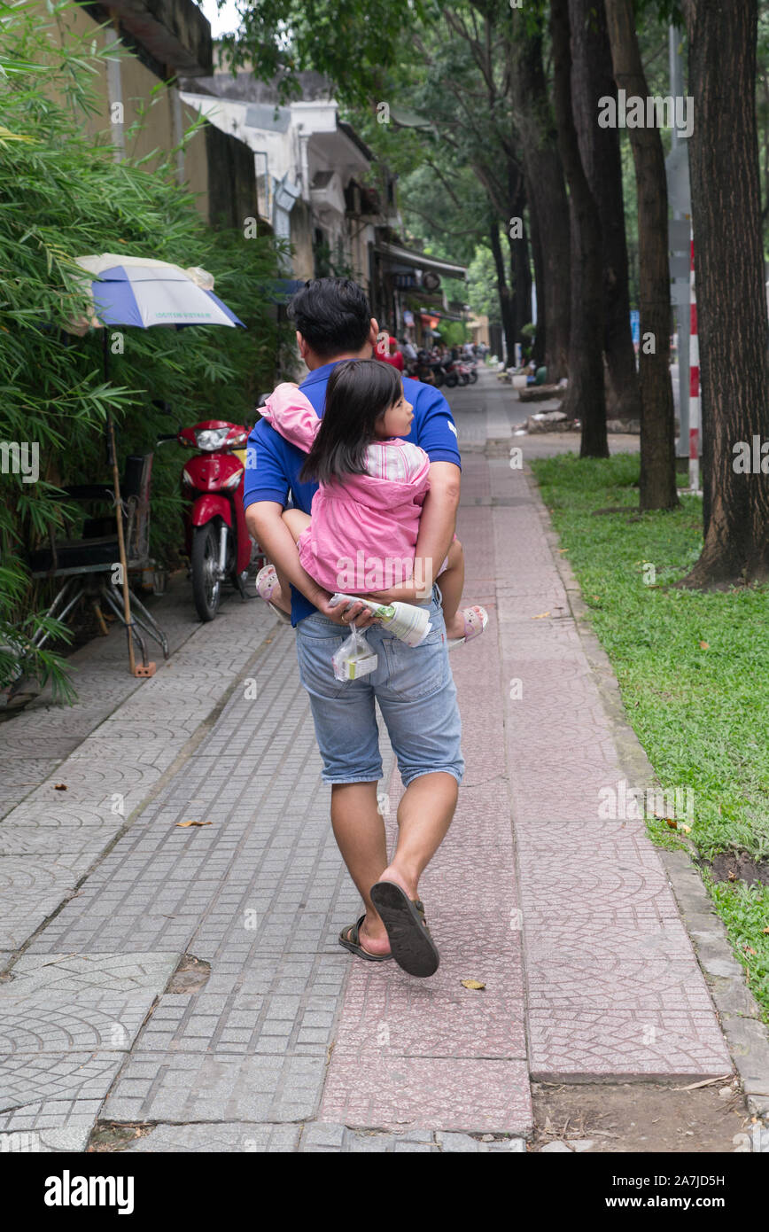 Father carrying petite fille sur le dos tout en descendant un trottoir bordé d'arbres sur Saigon journée d'été. Banque D'Images