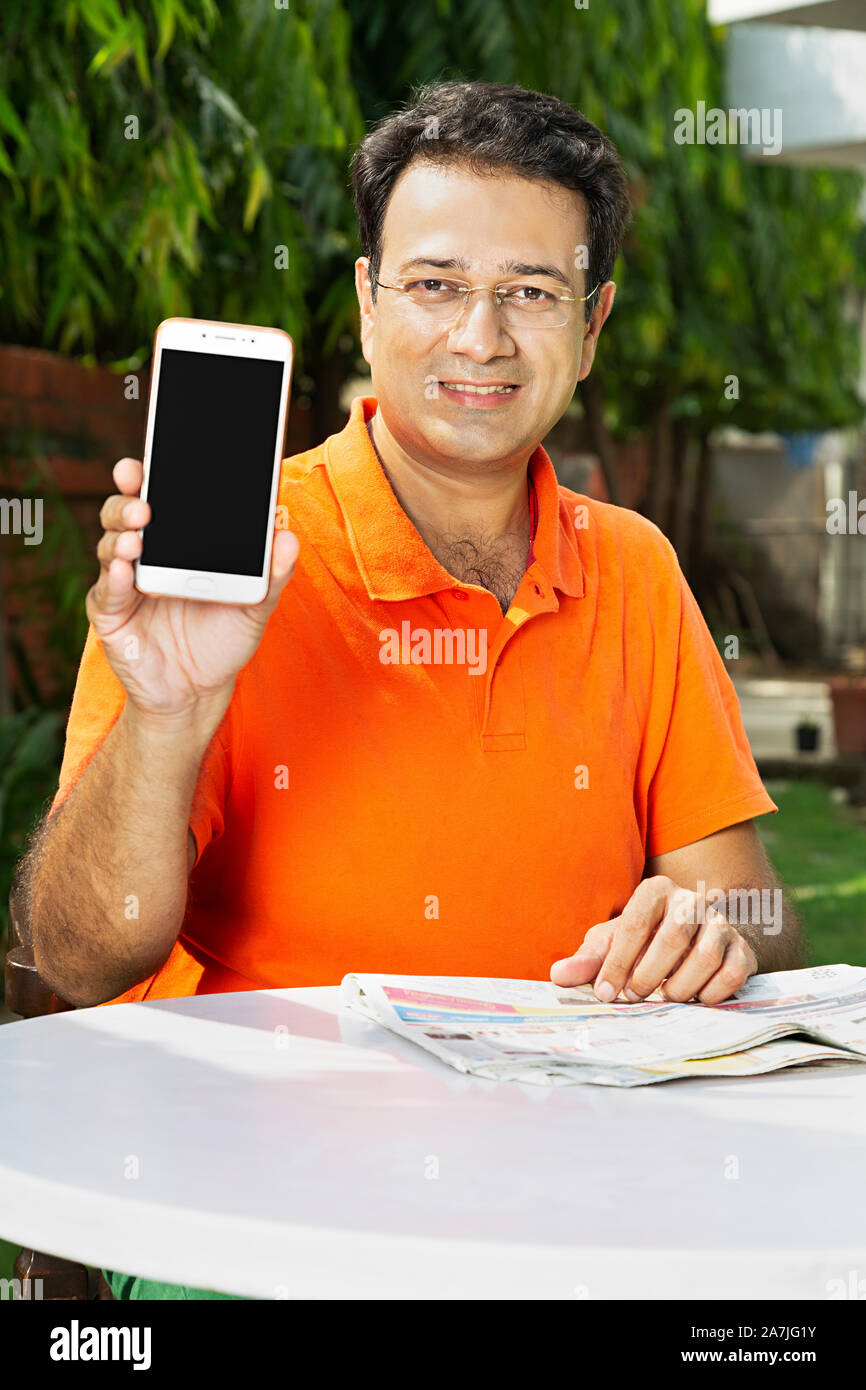 Portrait of Mid-Adult homme assis-sur-table tout en montrant téléphone cellulaire avec écran vide jardin In-Outdoor Banque D'Images