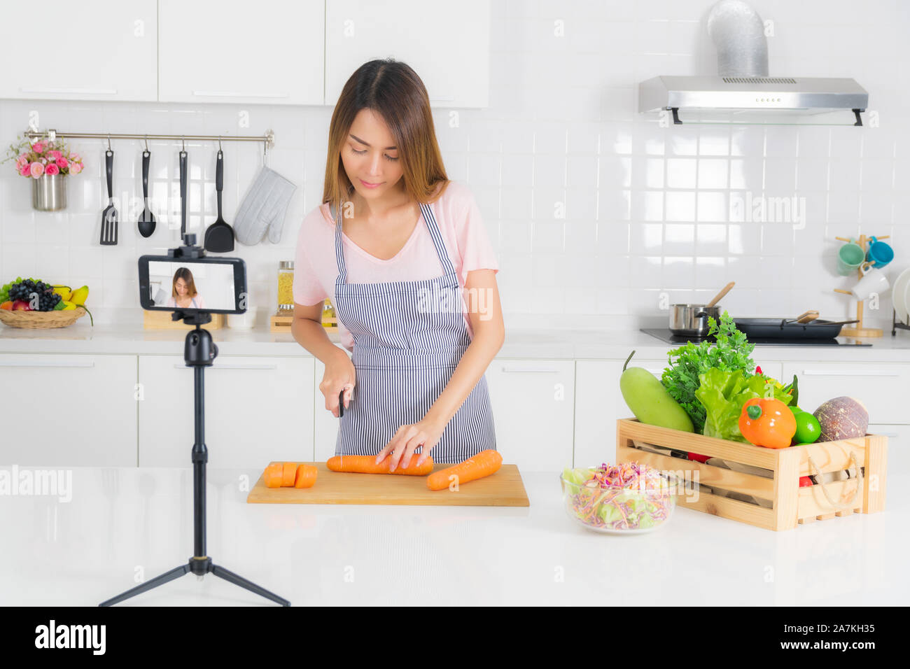 Belle femme asiatique, cheveux longs, portant un t-shirt rose à rayures et tablier, debout, trancher les carottes sur la table de cuisine moderne blanc et recordi Banque D'Images