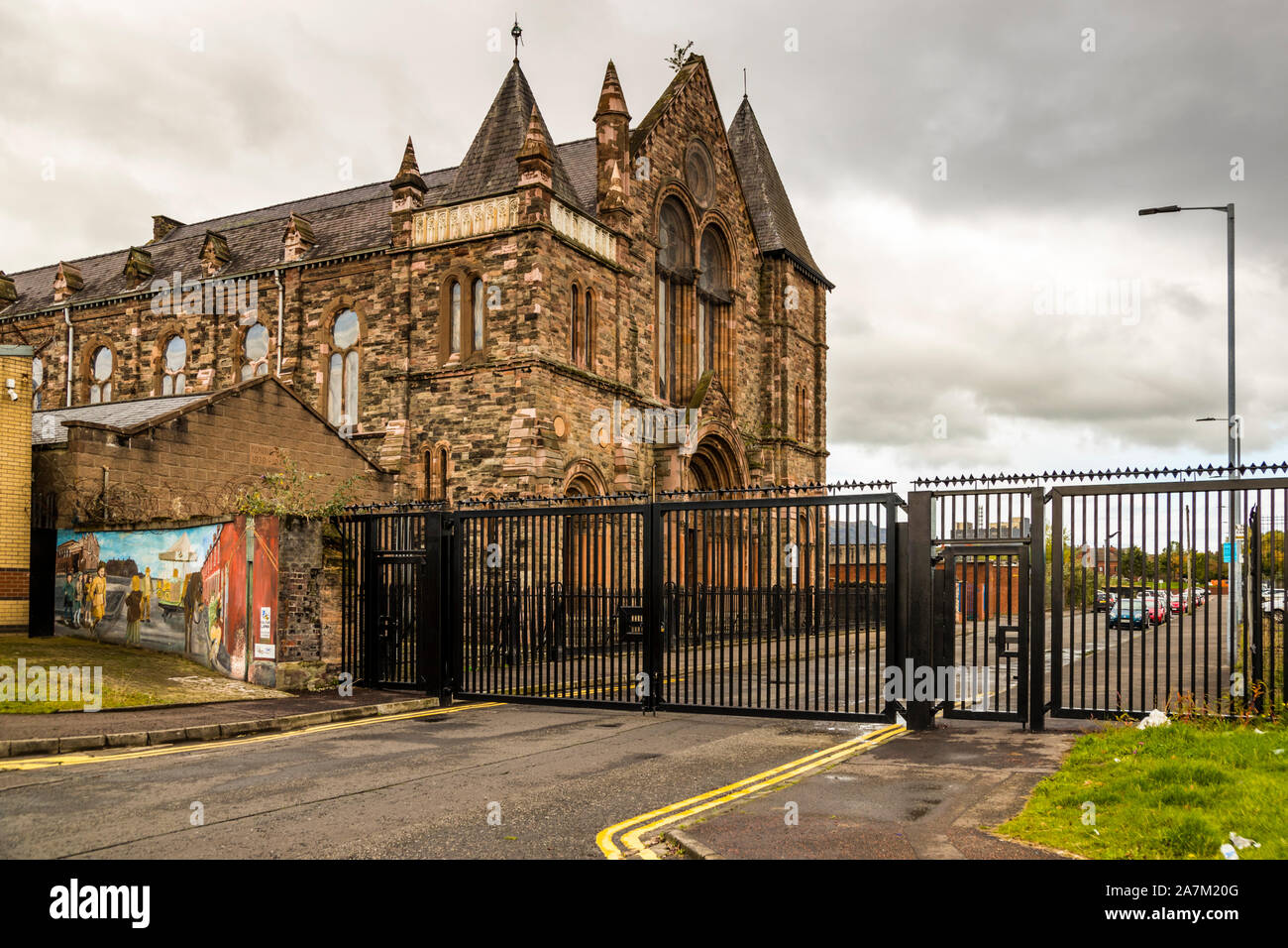 L'église presbytérienne de Townsend Street, construite en 1878 avec une façade néo-gothique en grès rouge à double tour, domine le coin de rue de Townsend Street, à Belfast, en Irlande du Nord ; une grille de sécurité noire surmontée de barbelés protège une ligne de paix qui passe par ici. À sa gauche, une petite fresque murale communautaire représentant une scène historique de rue est visible. Banque D'Images