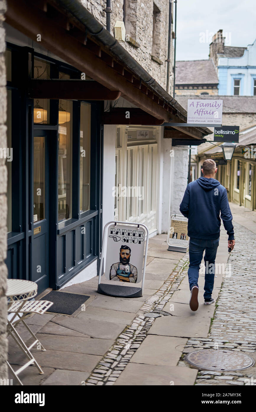 Homme passant devant un magasin de barbiers dans une rue ancienne et étroite de petits magasins indépendants à Kendal, Cumbria, Royaume-Uni, en bordure du Lake District Banque D'Images