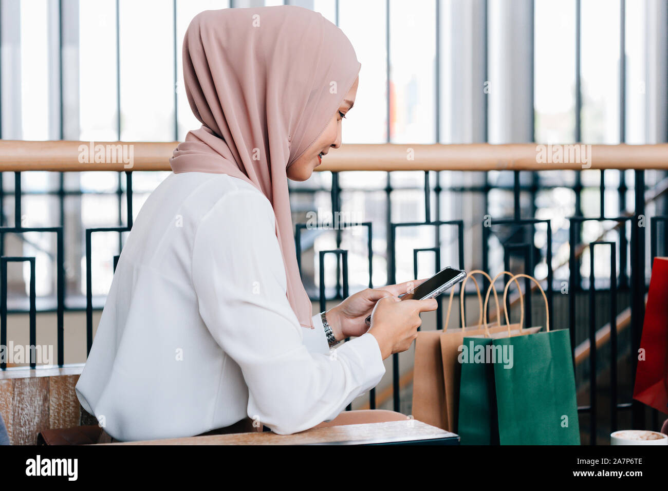 Vue de côté de madame asiatique en hijab smiling et la navigation mobile phone while sitting in cafe près de sacs en papier après le magasinage Banque D'Images
