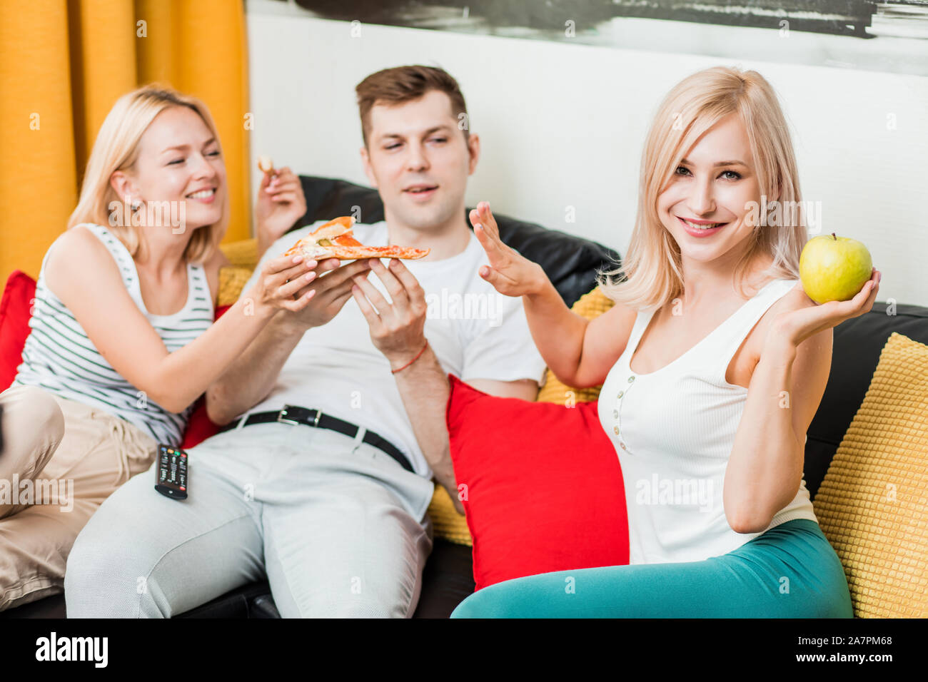 La malbouffe ou aliments sains. Belle sportive caucasienne blonde woman eating apple tout en profitant de ses amis pizza, faire stop, souriant à la recherche à Banque D'Images
