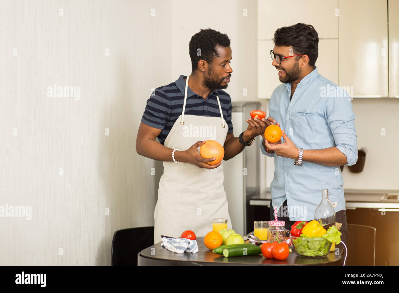 Deux beaux mâles joyeux amis debout dans la cuisine de fruits dans les mains, parler, faire du jus d'orange et de pamplemousse, la préparation des aliments sains t Banque D'Images