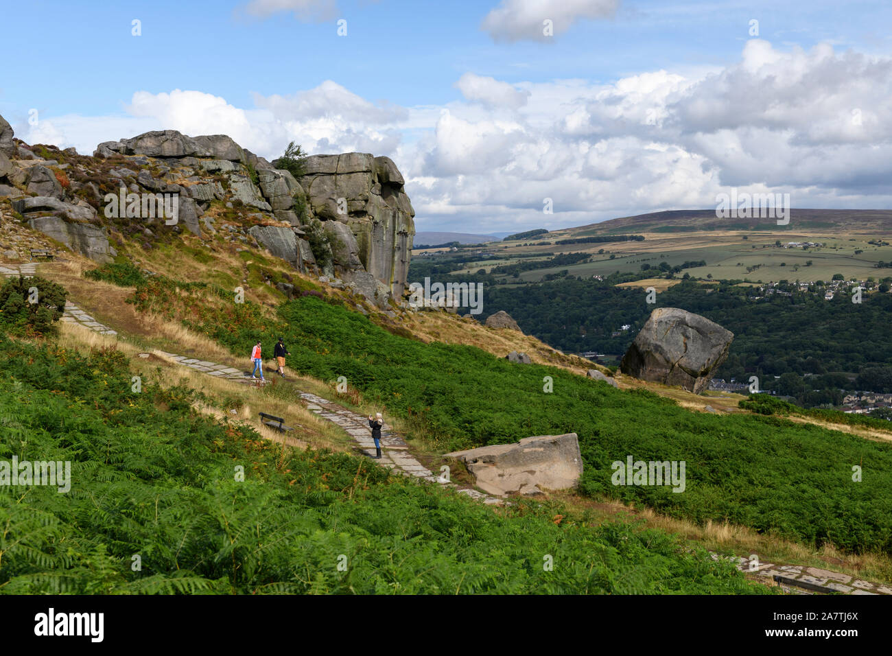 Les gens qui marchent sur le chemin par la haute vallée ensoleillée piton rocheux (ci-dessous) - paysage pittoresque de veau Vache et rochers, Ilkley Moor, West Yorkshire, Angleterre, Royaume-Uni. Banque D'Images
