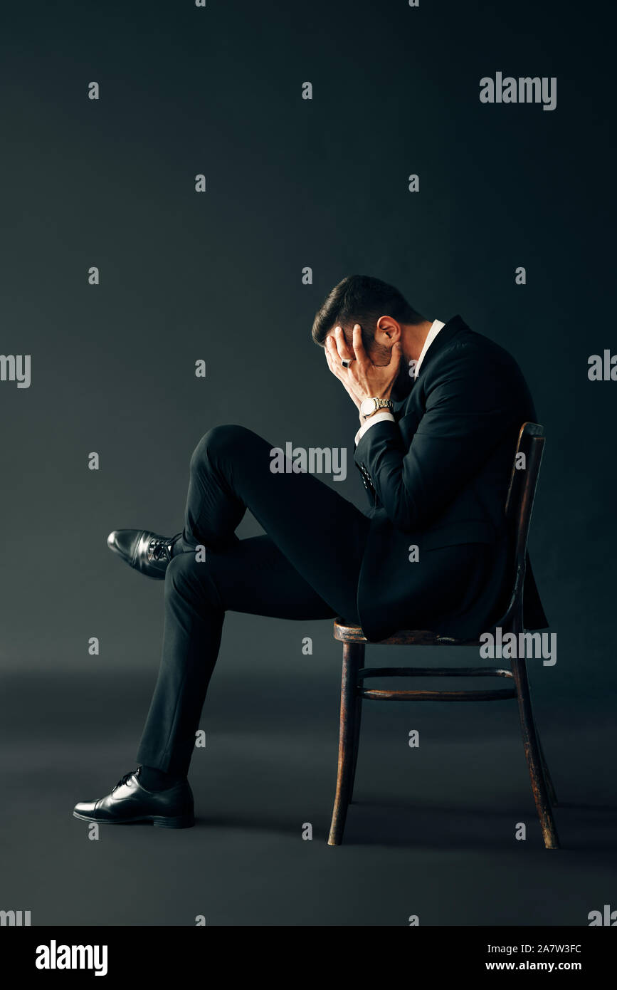 Triste Malheureux homme en costume noir assis sur la chaise et la face fermée par deux mains. Le profil Voir les portrait. La dépression et la douleur concept Banque D'Images