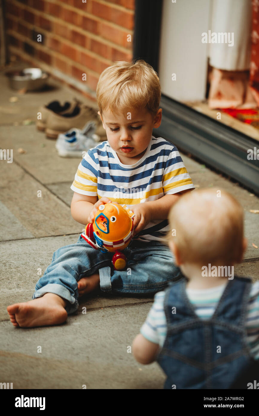 Tout-petit assis dehors jouer avec toy, regardée par frère bébé Banque D'Images