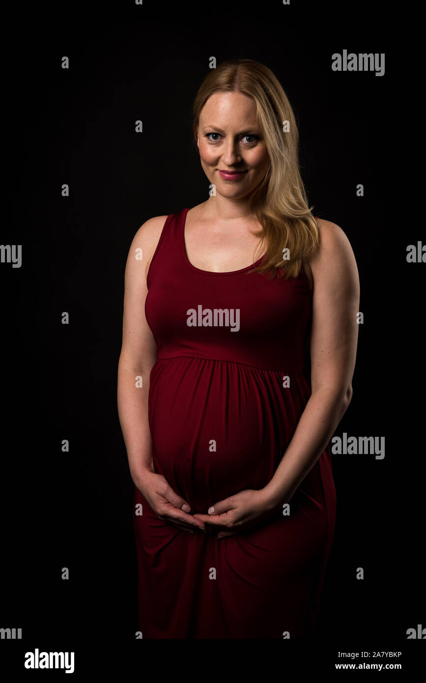 Femme enceinte en robe rouge holding belly sur fond noir. Portrait d'excité, heureux, blonde, jeune femme enceinte en studio photo. Concept de mat Banque D'Images