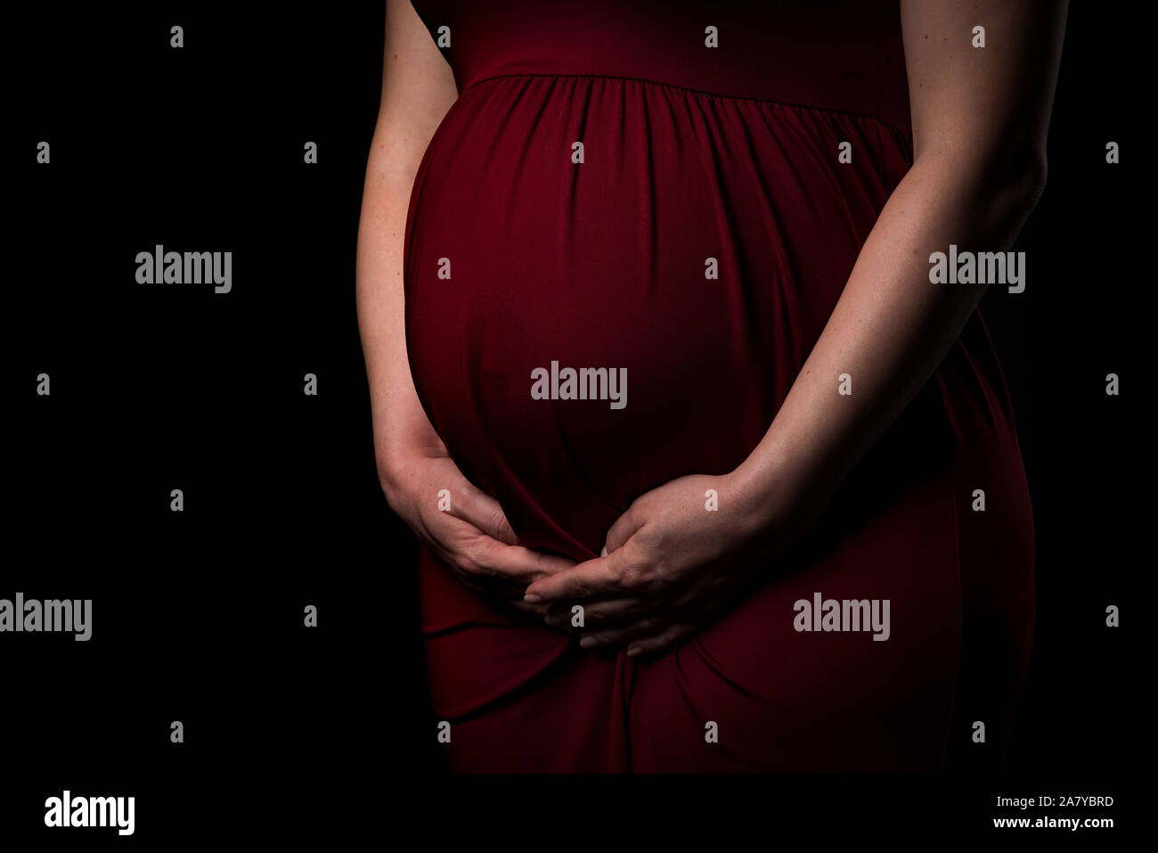 Femme enceinte en robe rouge holding belly sur fond noir. Portrait d'excité, heureux, blonde, jeune femme enceinte en studio photo. Concept de mat Banque D'Images