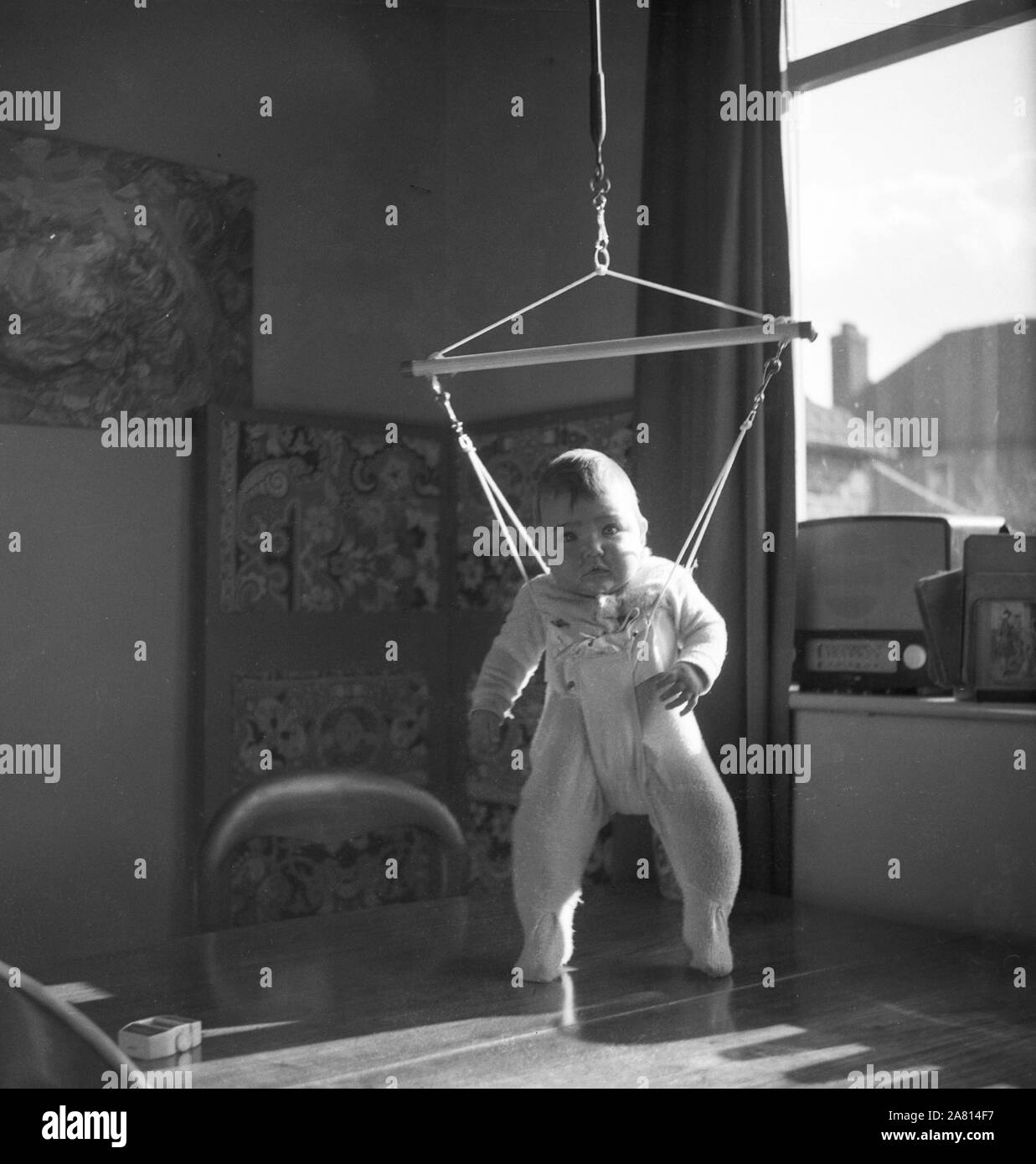 Années 1960, historique, à l'intérieur d'une chambre, un enfant bébé suspendu dans un harnais pivotant au-dessus d'une table, Angleterre, Royaume-Uni. Banque D'Images