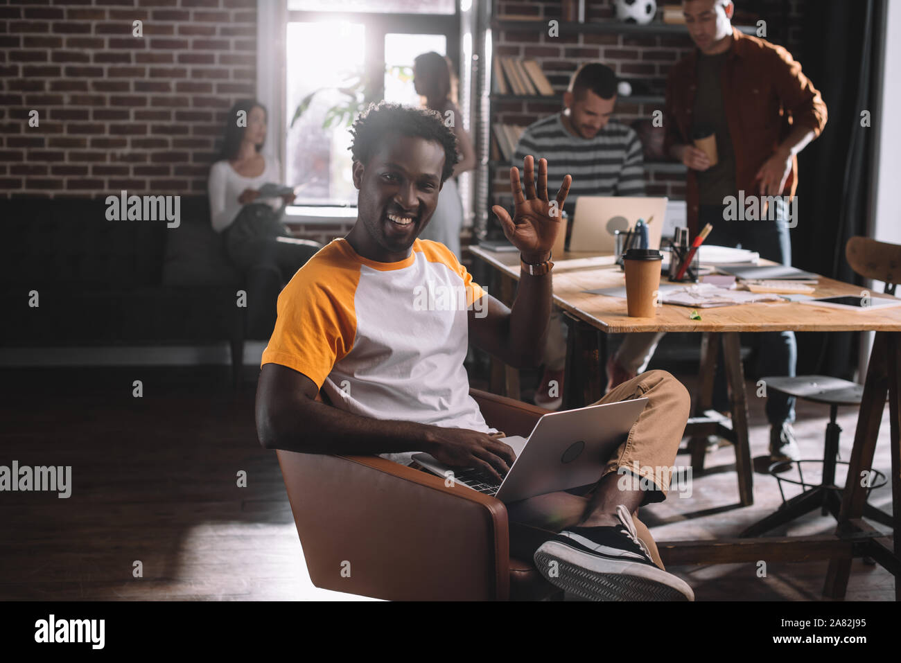 Smiling african american businessman using laptop while sitting in armchair et agitant la main près de jeunes collègues multiculturelle Banque D'Images