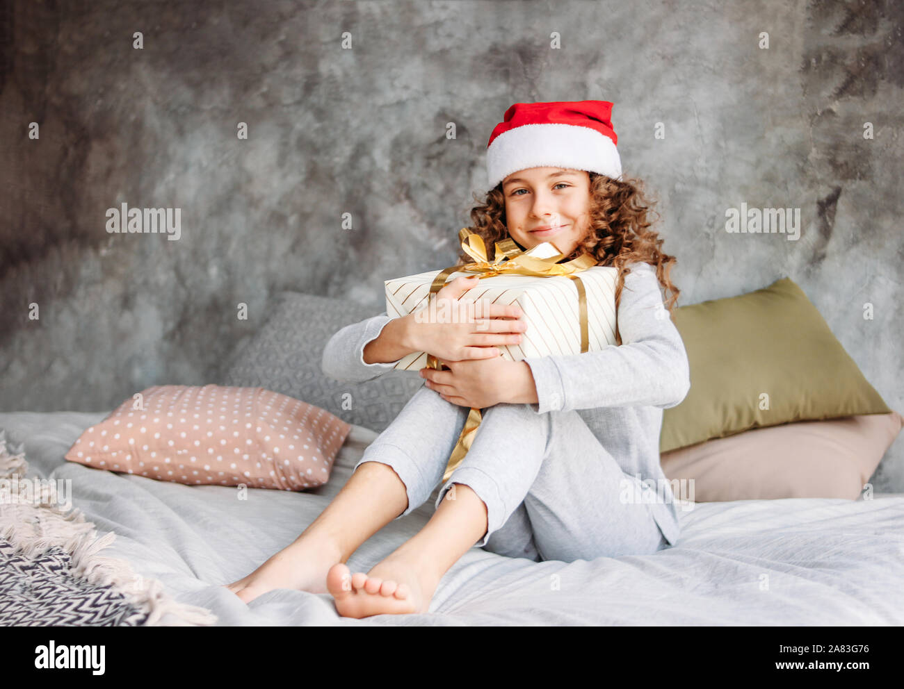 Belle jeune fille aux cheveux bouclés tween à Santa hat et pyjama assis sur le lit avec grande boîte-cadeau de Noël, temps matin Banque D'Images